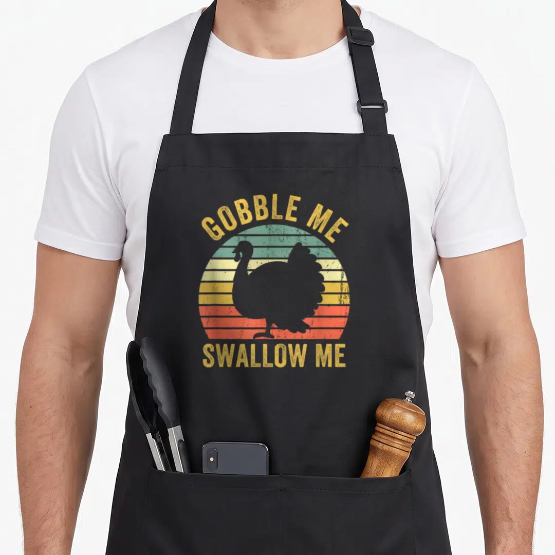 Swallow turkey apron - GOBBLE ME SWALLOW ME text over striped retro sun with grey turkey silhouette — shown on a black cotton canvas apron against a white background