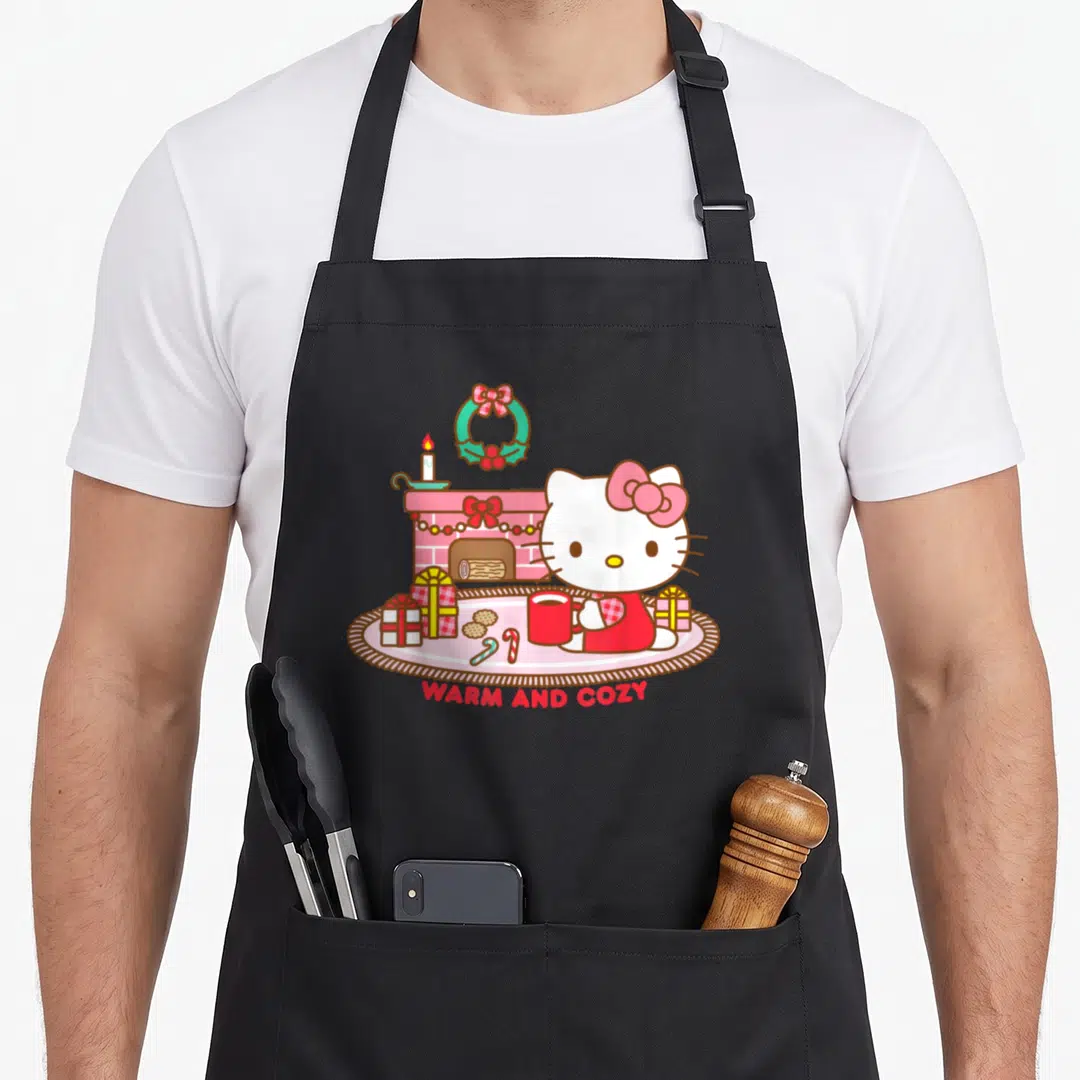 Warm christmas apron - Hello Kitty holding red mug near pink fireplace with WARM AND COZY text — shown on a black cotton canvas apron against a white background
