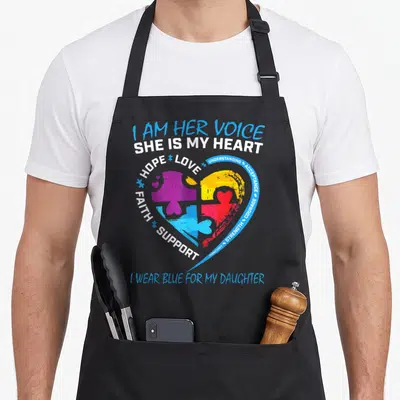 Puzzle mom daughter apron - Multi-colored puzzle piece heart with 'I AM HER VOICE' and 'I WEAR BLUE FOR MY DAUGHTER' — shown on a black cotton canvas apron against a white background