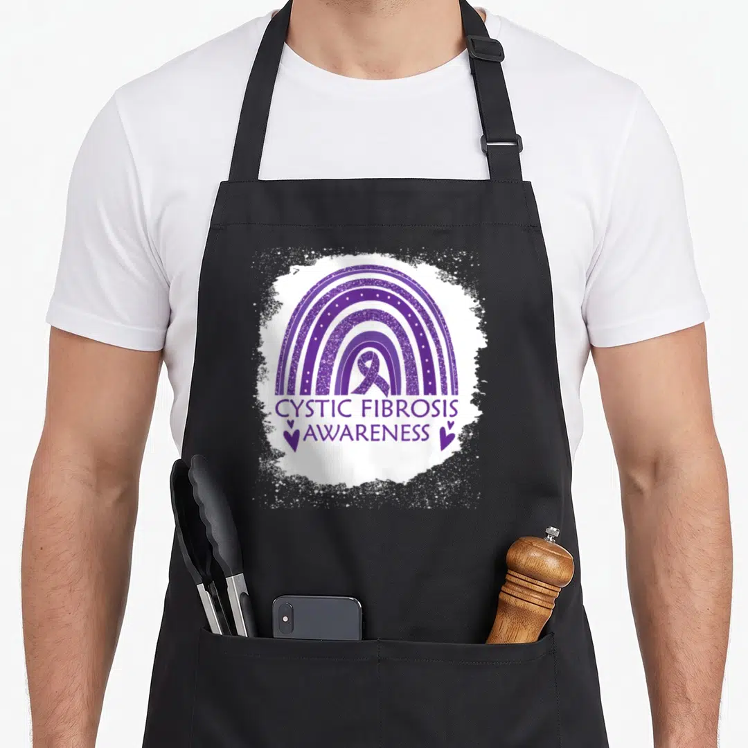 Cystic rainbow apron - Purple glittery rainbow with ribbon and text CYSTIC FIBROSIS AWARENESS flanked by hearts — shown on a black cotton canvas apron against a white background