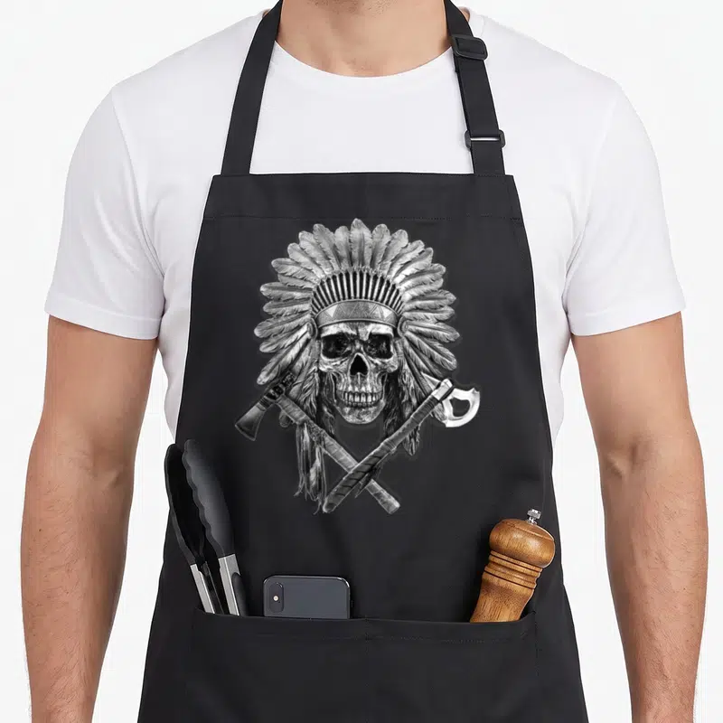 Pipe native american apron - Realistic skull with feathered headdress and two crossed tomahawks in silver grey and black — shown on a black cotton canvas apron against a white background