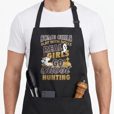 Paranormal hunter apron - Stacked brown and gold text with two white ghosts and a witch hat over a graveyard — shown on a black cotton canvas apron against a white background