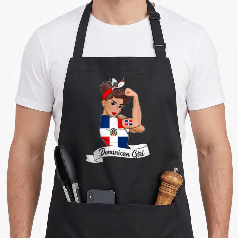 Dominicana pride apron - Cartoon woman in flag shirt flexing arm with Dominican Girl text on white ribbon — shown on a black cotton canvas apron against a white background