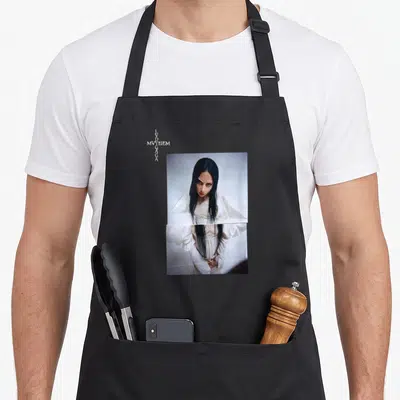 Mayhem novelty apron - Vertical LVD MVYHEM GAGA text beside a photo of a woman in a white dress — shown on a black cotton canvas apron against a white background