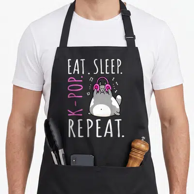 Sleep cat apron - Cartoon grey and white cat wearing pink headphones surrounded by Eat Sleep K Pop Repeat text — shown on a black cotton canvas apron against a white background