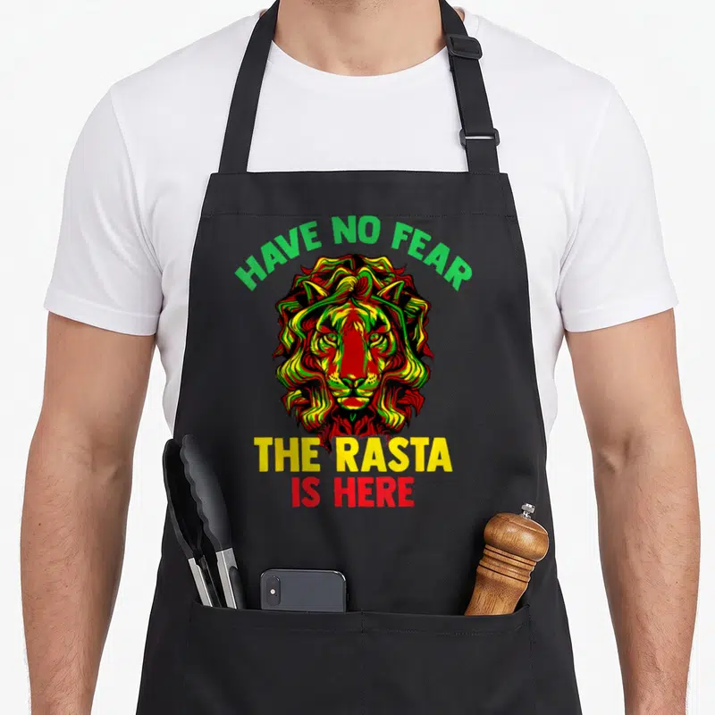 Raggea lion apron - Cartoon lion head with red yellow green black mane and text HAVE NO FEAR THE RASTA IS HERE — shown on a black cotton canvas apron against a white background