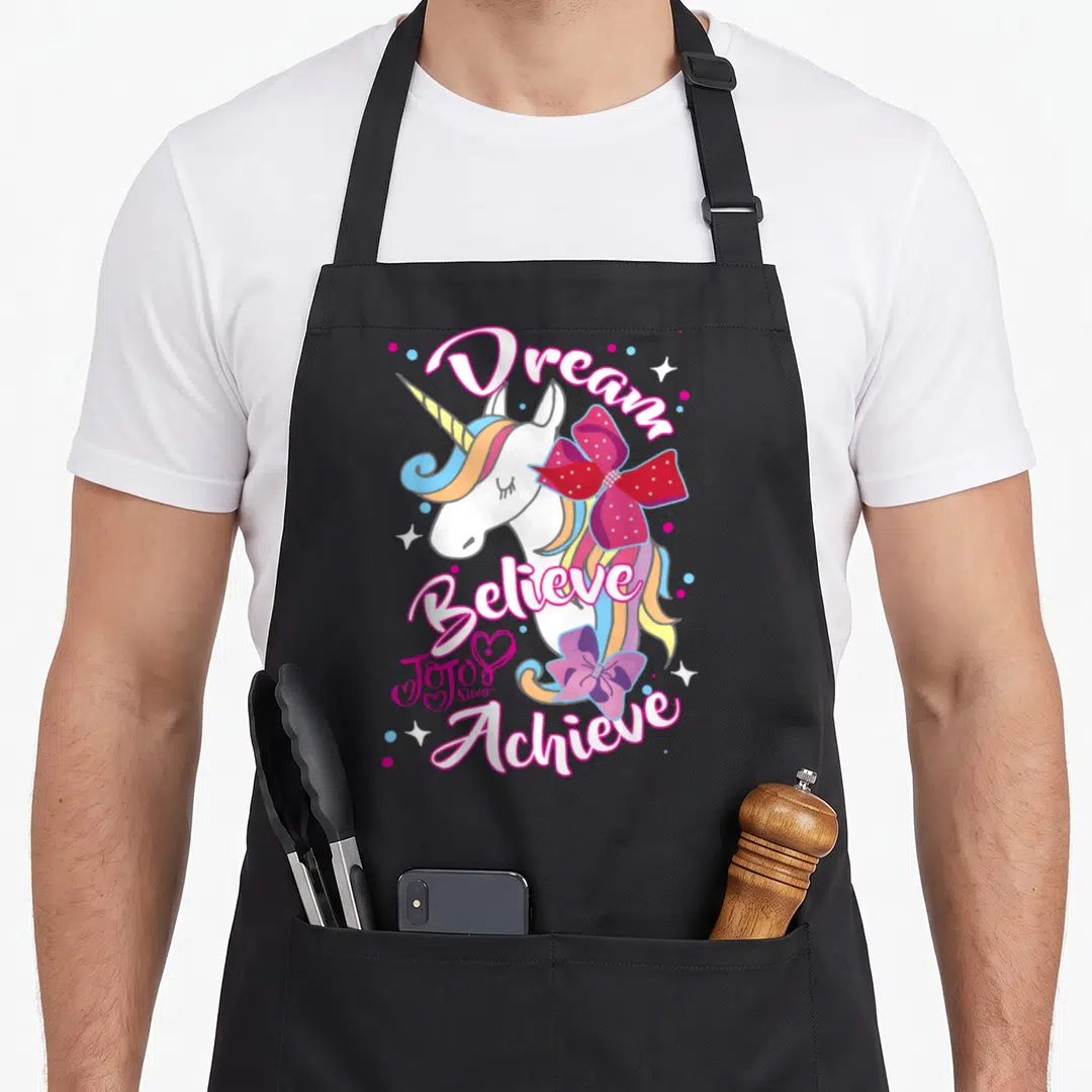 Dream unicorn apron - Cartoon unicorn with Dream Believe Achieve text, red and purple bows, rainbow mane — shown on a black cotton canvas apron against a white background