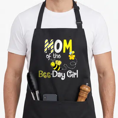 Birthday bee apron - MOM of the Bee-Day Girl text with two cartoon bees and dotted line — shown on a black cotton canvas apron against a white background
