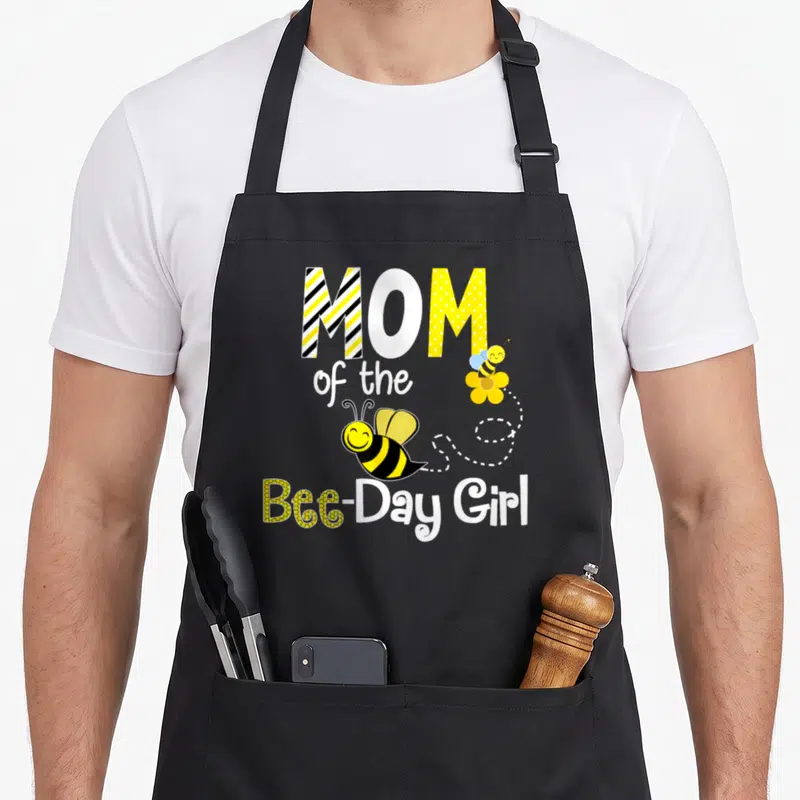 Birthday bee apron - MOM of the Bee-Day Girl text with two cartoon bees and dotted line — shown on a black cotton canvas apron against a white background