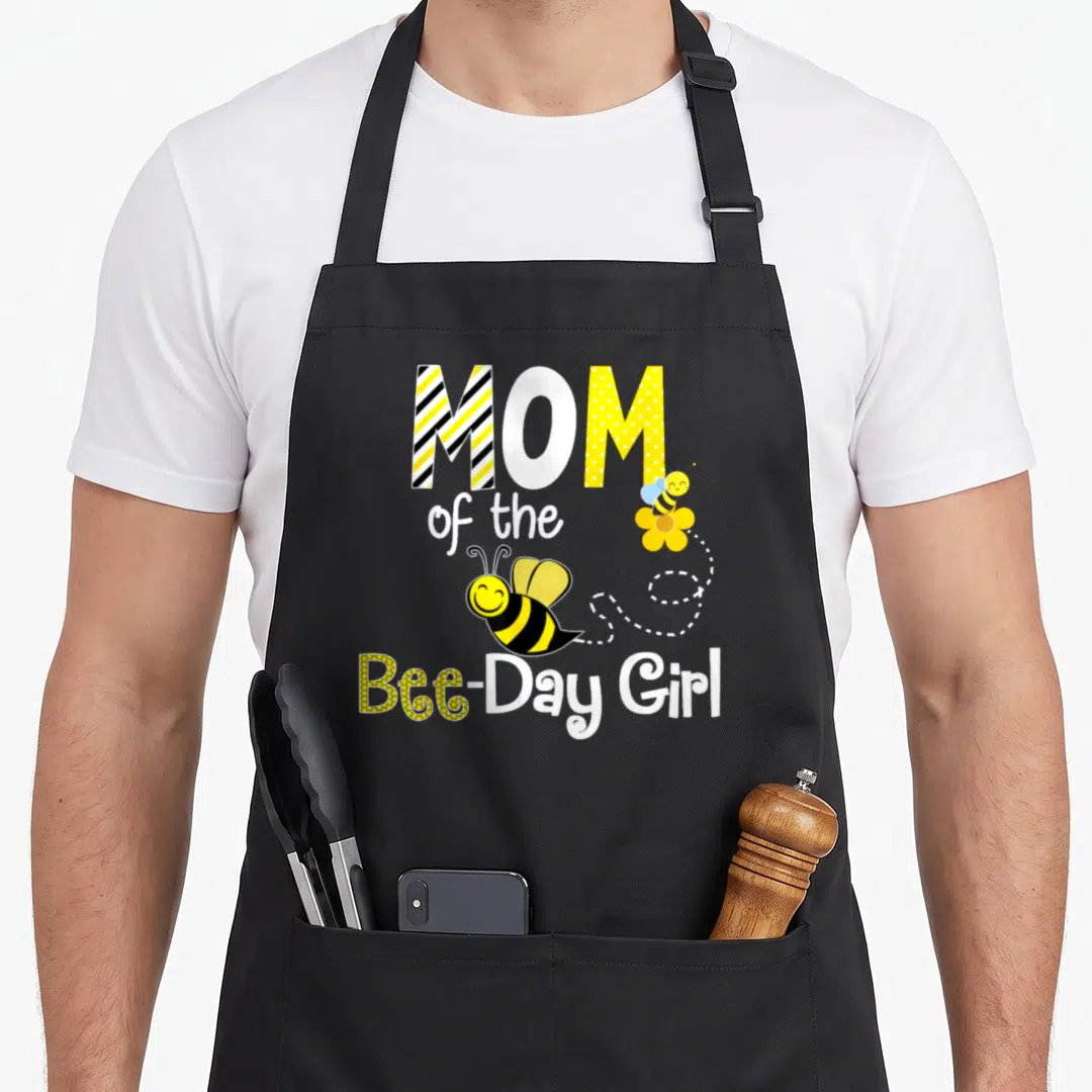 Birthday bee apron - MOM of the Bee-Day Girl text with two cartoon bees and dotted line — shown on a black cotton canvas apron against a white background