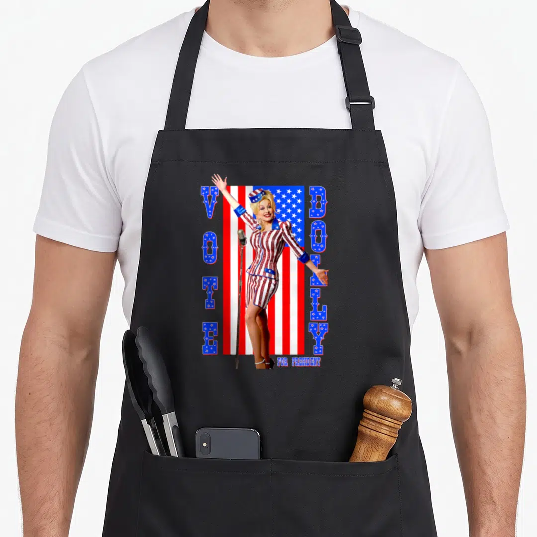 President dolly parton apron - Woman in patriotic suit with VOTE DOTLY FOR PRESIDENT text and USA flag background — shown on a black cotton canvas apron against a white background