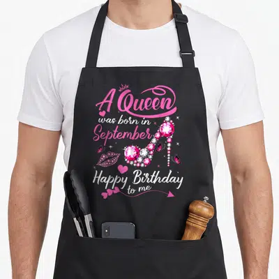 Funny queen september birthday apron - Jeweled pink high heel surrounded by text A Queen was born in September — shown on a black cotton canvas apron against a white background