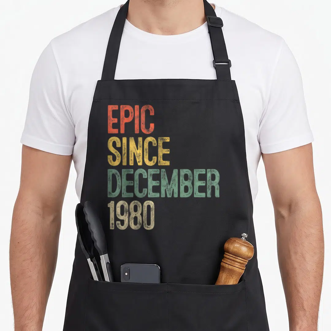 December 40th birthday apron - Stacked EPIC SINCE DECEMBER 1980 text in orange yellow green and beige retro typography — shown on a black cotton canvas apron against a white background