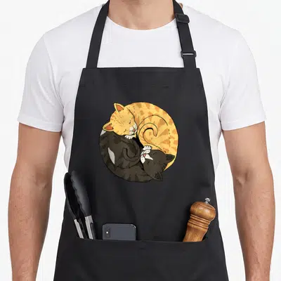 Yang cat apron - Two cats curled in yin yang pose, orange and black with white accents, cartoon style — shown on a black cotton canvas apron against a white background