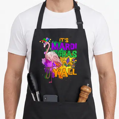 Carnival mardi gras aprons - Pink flamingo in jester hat holding beads beside IT'S MARDI GRAS Y'ALL text — shown on a black cotton canvas apron against a white background