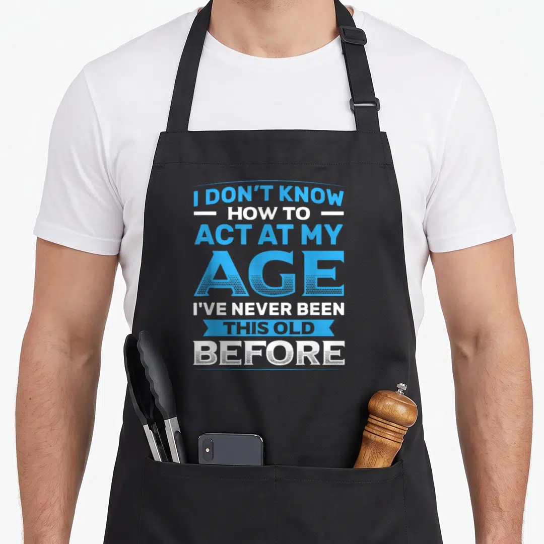 Act birthday apron - Stacked blue and white text reading I DON'T KNOW HOW TO ACT AT MY AGE — shown on a black cotton canvas apron against a white background