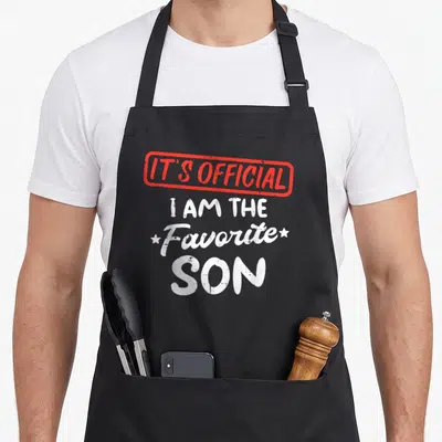 Youth family aprons - Red stamp border with IT'S OFFICIAL text above white I AM THE Favorite SON lettering flanked — shown on a black cotton canvas apron against a white background