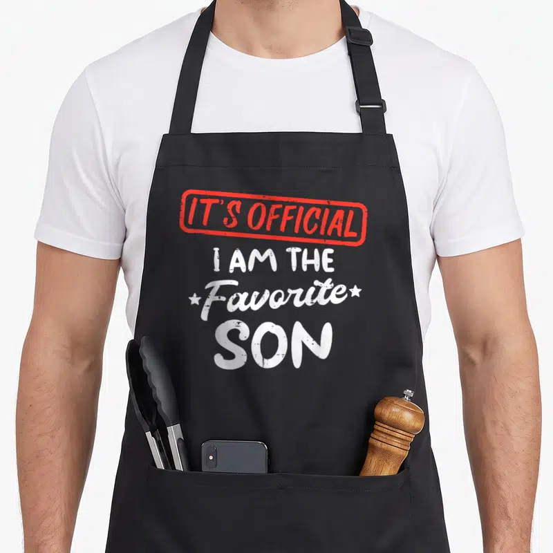 Youth family aprons - Red stamp border with IT'S OFFICIAL text above white I AM THE Favorite SON lettering flanked — shown on a black cotton canvas apron against a white background