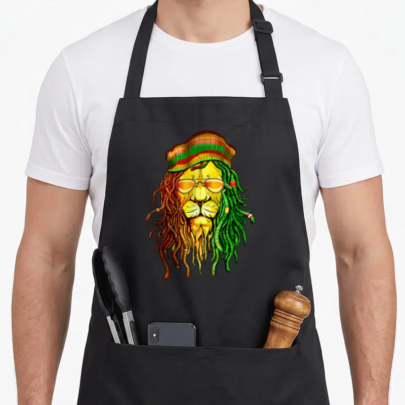 Reggae lion apron - Cartoon lion head with red yellow green dreadlocks and striped beanie — shown on a black cotton canvas apron against a white background