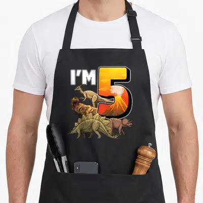 Birthday 5th dinosaur apron - I'M 5 text with volcano inside number five and four cartoon dinosaurs in orange and brown — shown on a black cotton canvas apron against a white background