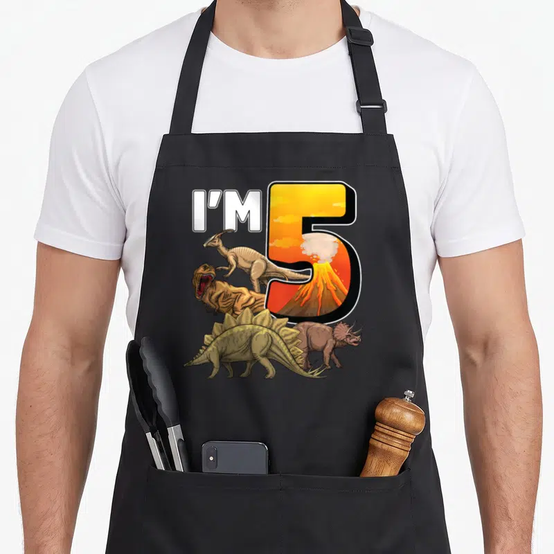 Birthday 5th dinosaur apron - I'M 5 text with volcano inside number five and four cartoon dinosaurs in orange and brown — shown on a black cotton canvas apron against a white background