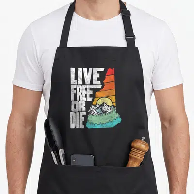 Free apron or apron - LIVE FREE OR DIE text next to NH outline filled with retro sunset and mountains — shown on a black cotton canvas apron against a white background