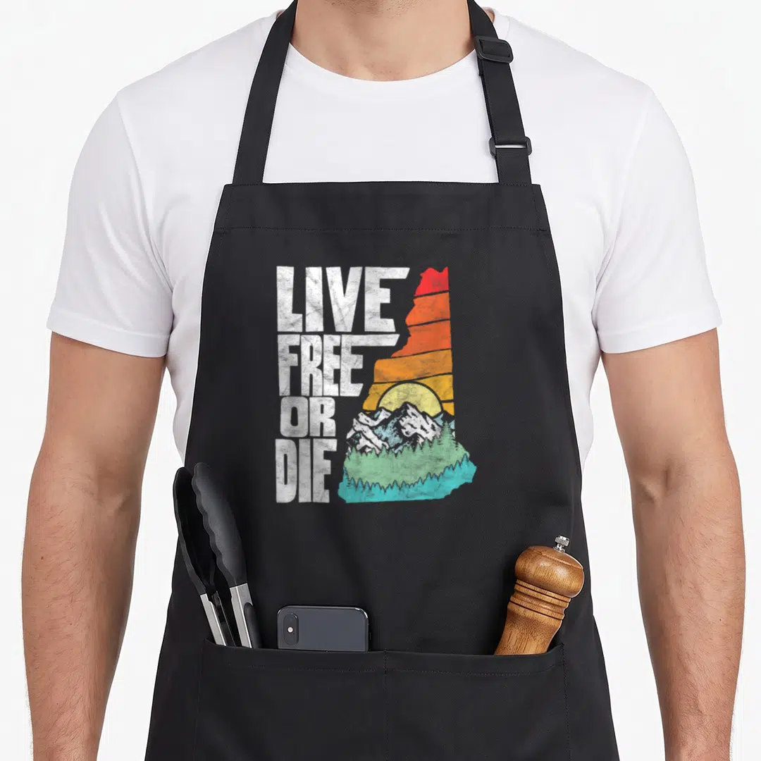 Free apron or apron - LIVE FREE OR DIE text next to NH outline filled with retro sunset and mountains — shown on a black cotton canvas apron against a white background