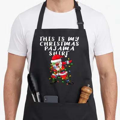 Pajama christmas santa apron - Santa Claus dabbing while tangled in Christmas lights under white stacked text — shown on a black cotton canvas apron against a white background