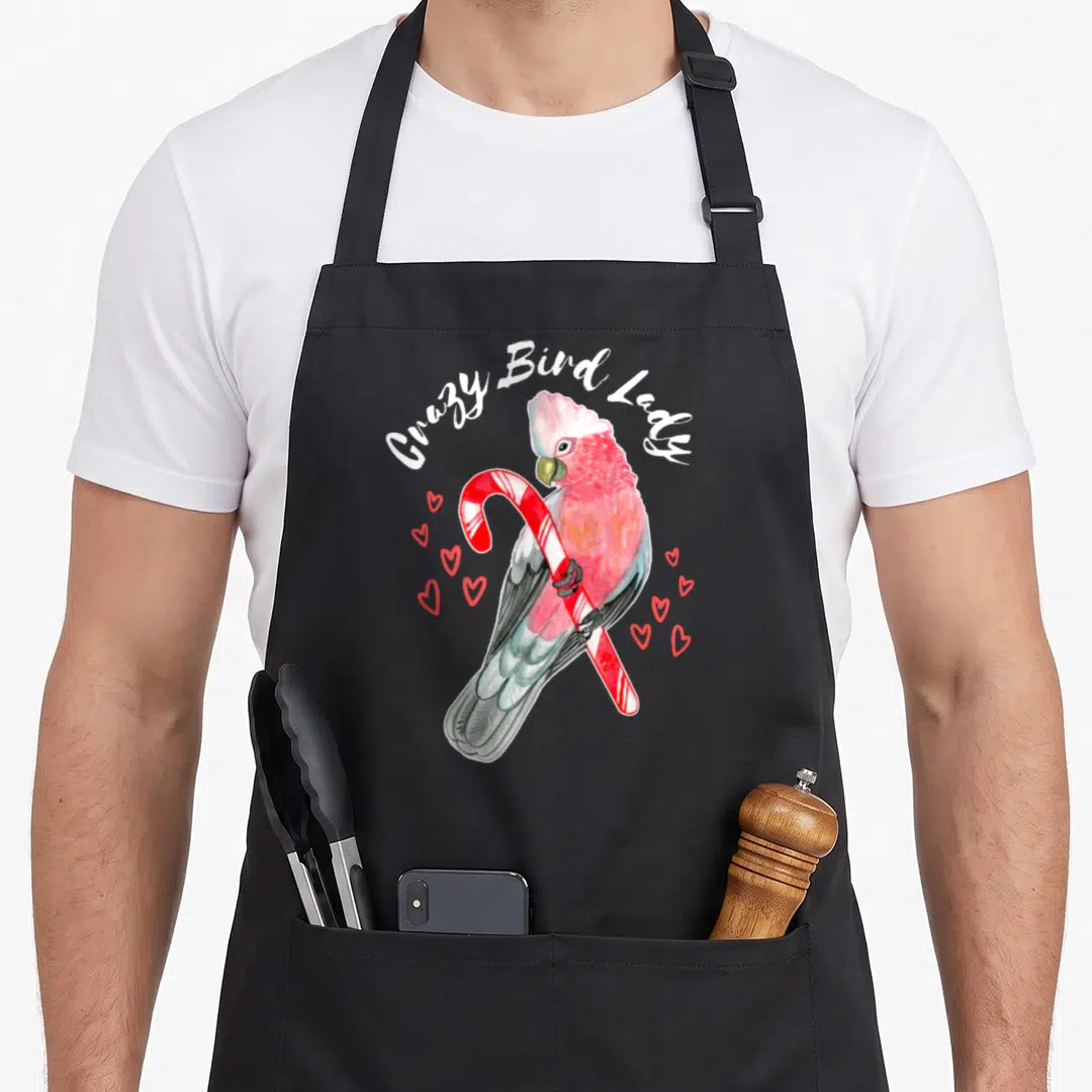 Rose bird apron - Cockatoo holding candy cane surrounded by red hearts with arched text in watercolor style — shown on a black cotton canvas apron against a white background