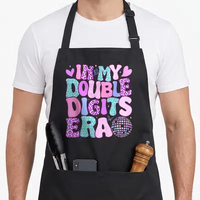 Digit double apron - Stacked IM MY DOUBLE DIGITS ERA text with disco ball, hearts, and butterflies in pink blue purple — shown on a black cotton canvas apron against a white background