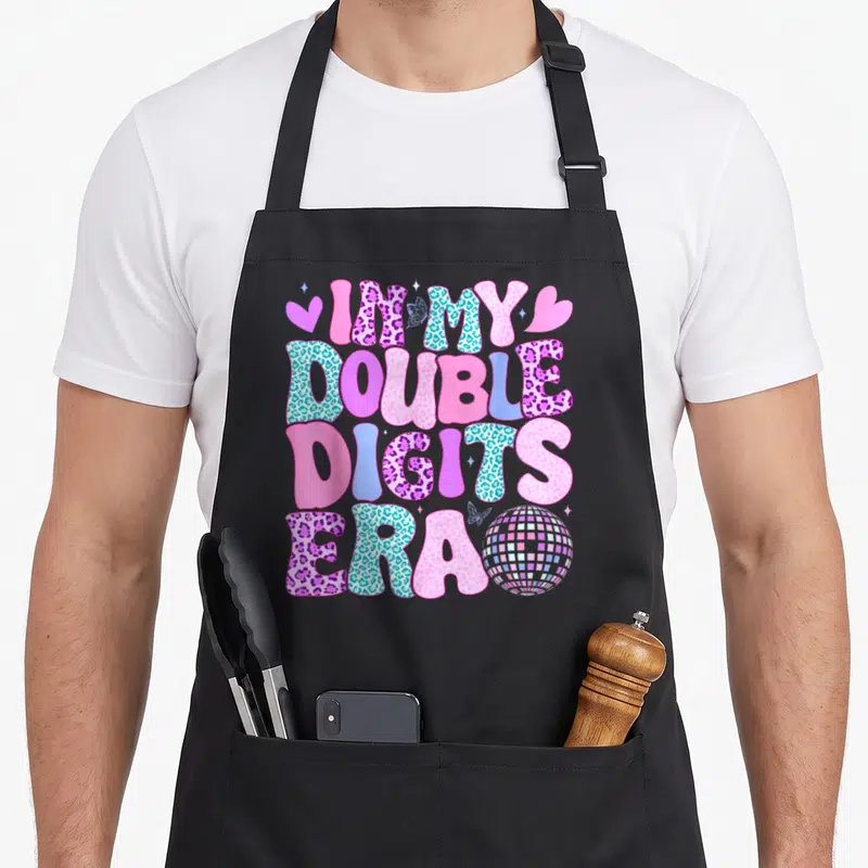 Digit double apron - Stacked IM MY DOUBLE DIGITS ERA text with disco ball, hearts, and butterflies in pink blue purple — shown on a black cotton canvas apron against a white background