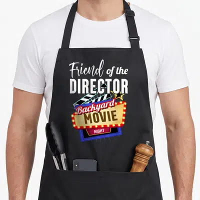 Backyard friends apron - Friend of the Director text with marquee sign and clapperboard — shown on a black cotton canvas apron against a white background
