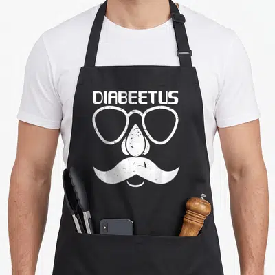 Diabete beard apron - White distressed DIABEETUS text above glasses nose and mustache forming face — shown on a black cotton canvas apron against a white background