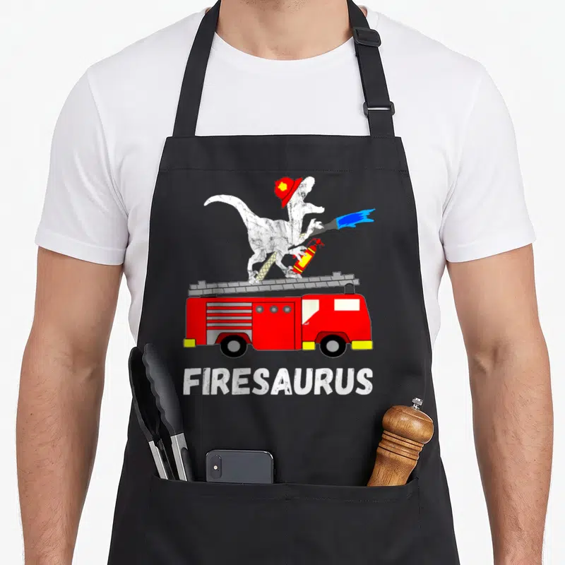 Fire dinosaur apron - White dinosaur in red hat on red fire truck spraying blue water with FIRESAURUS text below — shown on a black cotton canvas apron against a white background