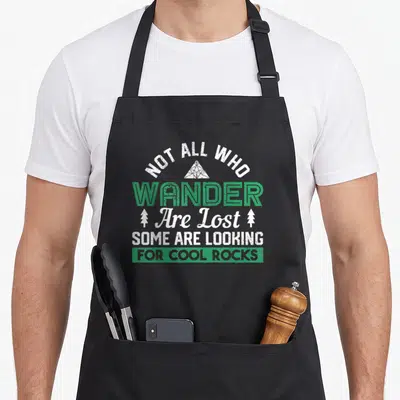 Geologist hunter apron - Stacked text reading Not All Who Wander Are Lost Looking For Cool Rocks with triangle and trees — shown on a black cotton canvas apron against a white background