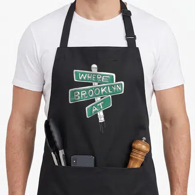 Brooklyn apron for women - Green street sign graphic with white text reading WHERE BROOKLYN AT stacked vertically — shown on a black cotton canvas apron against a white background