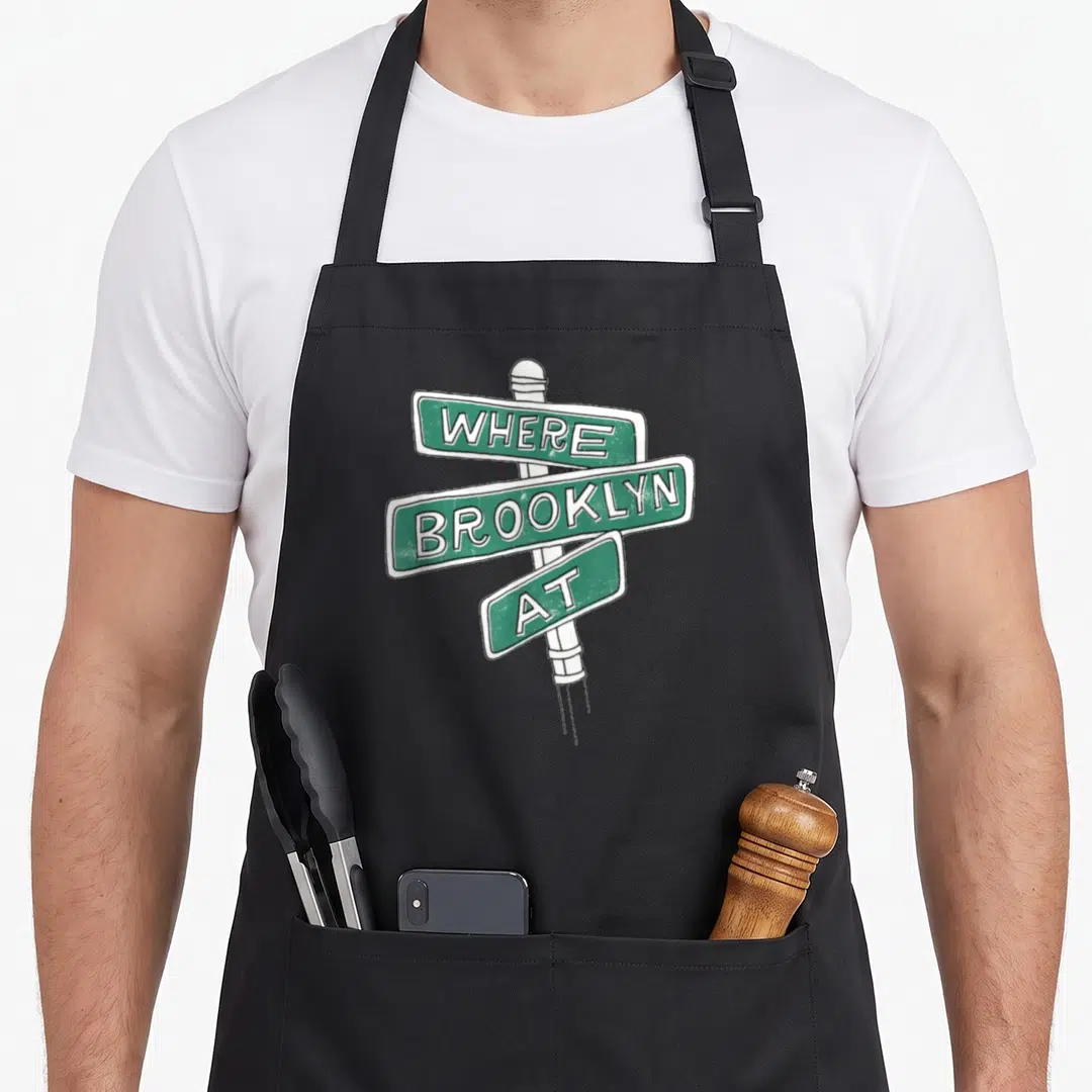 Brooklyn apron for women - Green street sign graphic with white text reading WHERE BROOKLYN AT stacked vertically — shown on a black cotton canvas apron against a white background