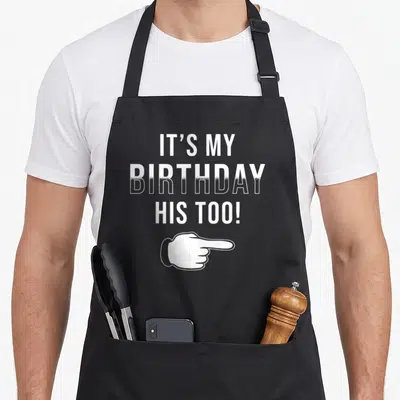 Twin his birthday apron - White stacked text It's My Birthday His Too! with pointing hand on black bib — shown on a black cotton canvas apron against a white background