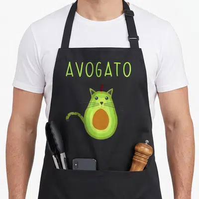 Avogato cat apron - AVOGATO text above cartoon avocado-cat with brown pit, red stem, and striped tail — shown on a black cotton canvas apron against a white background