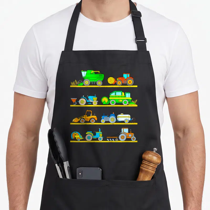 Combine vehicle apron - Eight cartoon farm vehicles on four yellow shelves in green orange blue and yellow — shown on a black cotton canvas apron against a white background