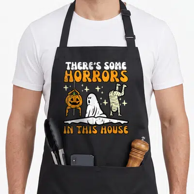Horror halloween apron - Cartoon pumpkin ghost and mummy below text There's Some Horrors In This House — shown on a black cotton canvas apron against a white background