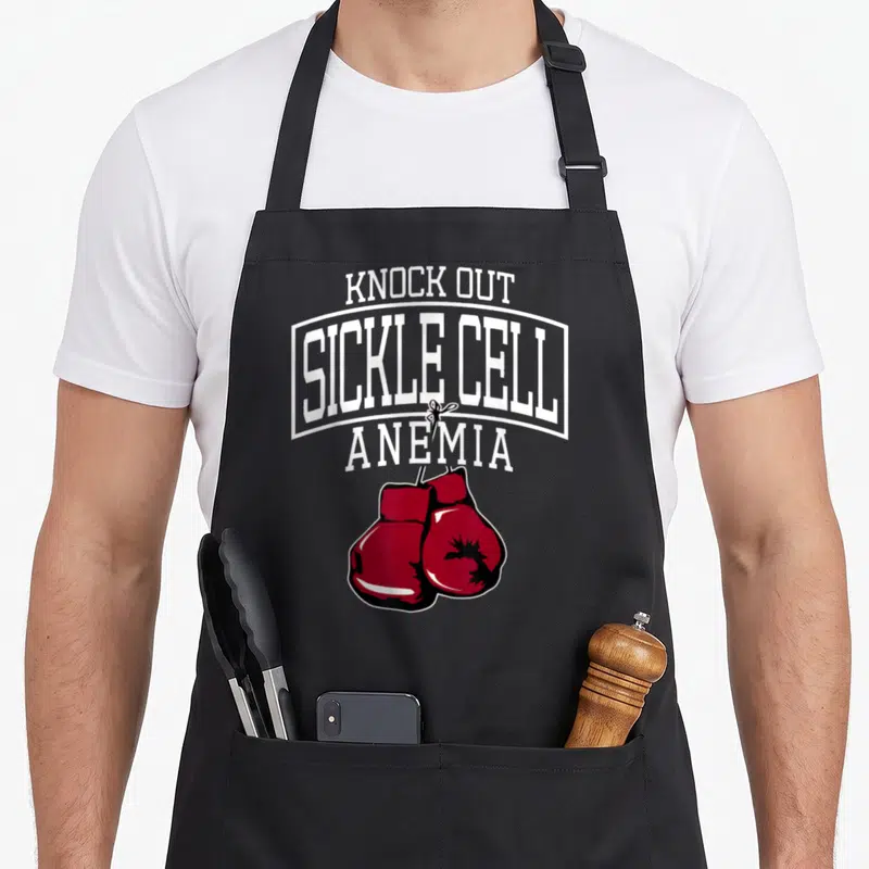 Support sickle cell awareness apron - Stacked white text reading Knock Out Sickle Cell Anemia above a pair of red — shown on a black cotton canvas apron against a white background