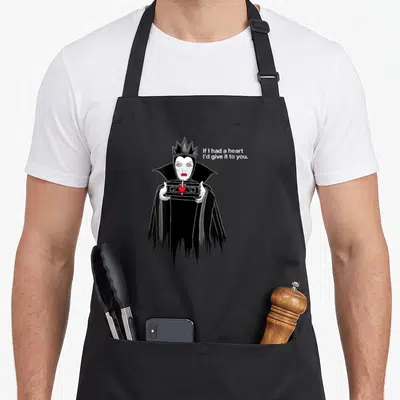 Evil queen apron - Cartoon queen holding red heart box with white sarcastic text beside her head — shown on a black cotton canvas apron against a white background