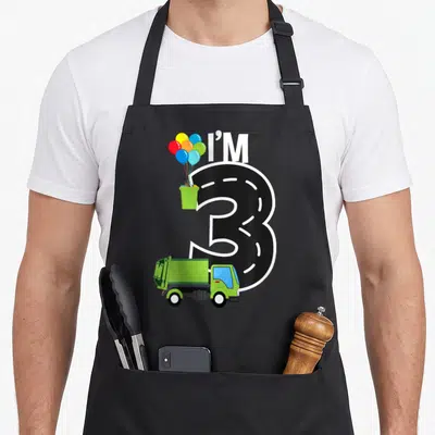 Three birthday truck apron - I'M 3 road number with green garbage truck and balloons in trash can — shown on a black cotton canvas apron against a white background