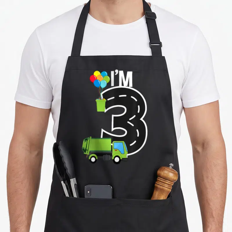 Three birthday truck apron - I'M 3 road number with green garbage truck and balloons in trash can — shown on a black cotton canvas apron against a white background