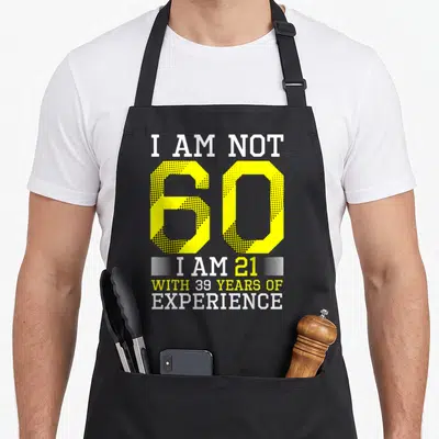 Cool 60th birthday apron - Yellow 60 typography saying I am not 60 I am 21 with 39 years experience — shown on a black cotton canvas apron against a white background