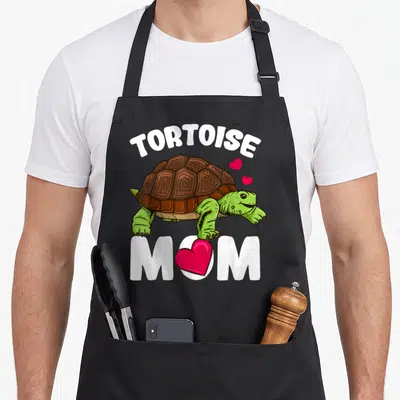 Funny tortoise apron - Cartoon tortoise in green and brown with white text and pink hearts replacing the O in MOM — shown on a black cotton canvas apron against a white background