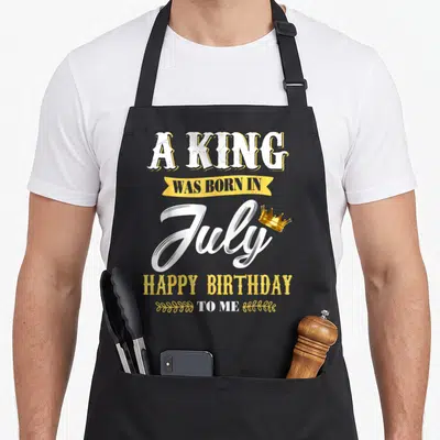 July birthday king apron - Stacked white and yellow text A KING WAS BORN IN July HAPPY BIRTHDAY TO ME with gold crown — shown on a black cotton canvas apron against a white background