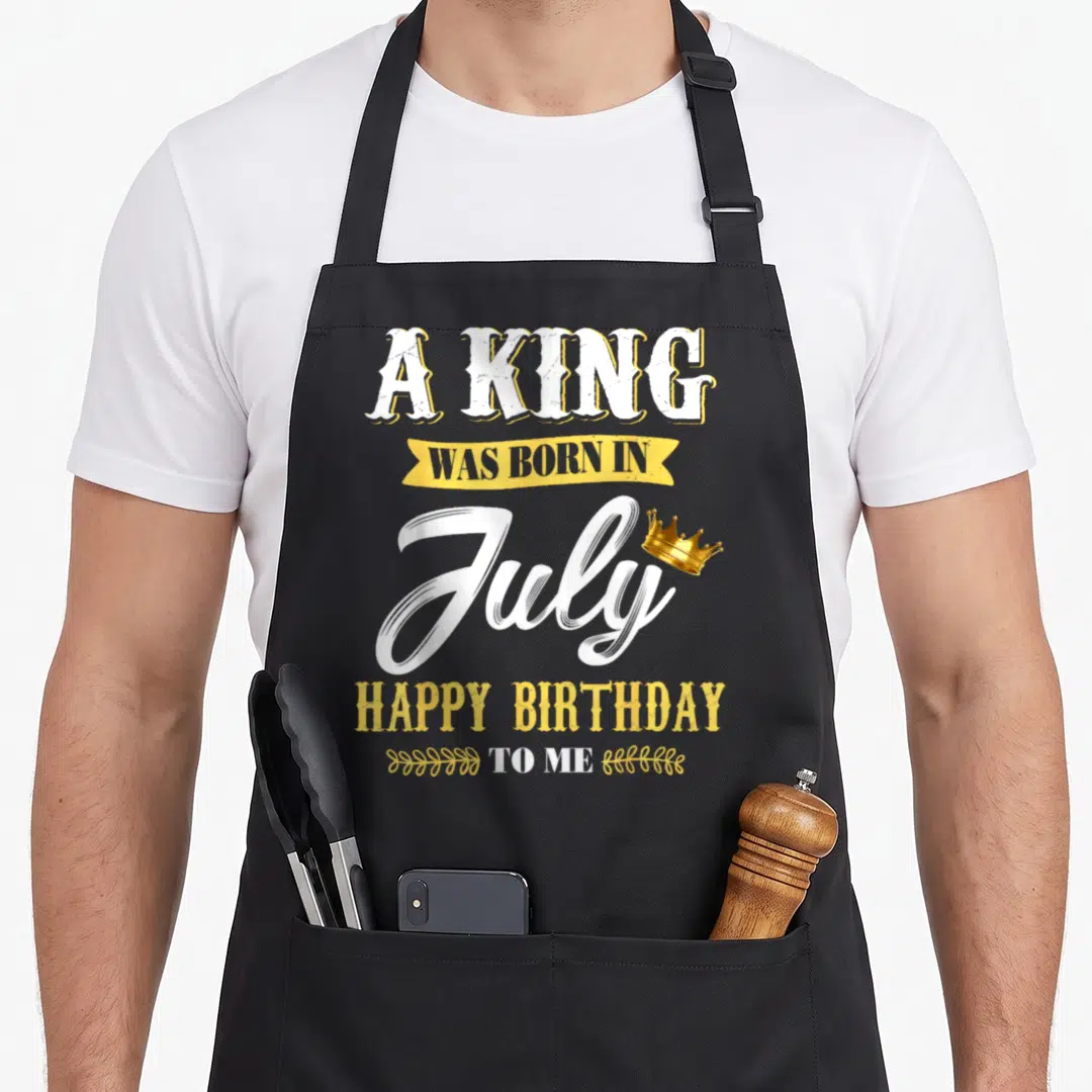 July birthday king apron - Stacked white and yellow text A KING WAS BORN IN July HAPPY BIRTHDAY TO ME with gold crown — shown on a black cotton canvas apron against a white background