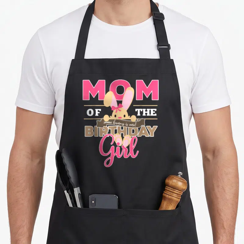 Funny birthday bunny apron - Cartoon bunny overlapping Mom of the Birthday Girl text with brown banner reading some — shown on a black cotton canvas apron against a white background